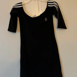 Svart klänning från Adidas - Snygg svart klänning från Adidas med trekvartsärm och klassiska vita ränder på axlarna. Perfekt för en sportig och stilren look. Adidas-loggan är diskret placerad på bröstet.