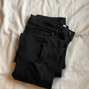 Svarta jeans med slitningar - Snygga svarta jeans med slitningar på benen. De har en klassisk design med knappar och dragkedja. Perfekta för en trendig look. Använd två gånger 