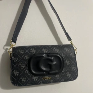 Svart axelväska från Guess - Snygg svart axelväska från Guess med logotypmönster och en stor G-detalj på framsidan. Perfekt för att bära dina nödvändigheter med stil. Helt ny aldrig använd.