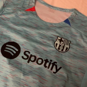 Ljusblå/turkos fotbollströja med FCB-logga🩵💚 - Snygg ljusblå fotbollströja med FCB-logga och sponsortryck från Spotify. Tröjan har ett unikt mönster i rött och blått samt spelartryck med namnet Lewandowski och nummer 9 på ryggen. Perfekt för fotbollsfans! ⚠️Ej äkta⚠️Helt oanvänd❤️❤️