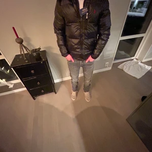 Svart dunjacka från Polo Ralph Lauren - Snygg svart dunjacka från Polo Ralph Lauren med röd logga på bröstet. Jackan har dragkedja framtill och flera praktiska fickor. Perfekt för kyliga dagar med sin vadderade design och avtagbar luva. Den är i storlek s passar 155-175