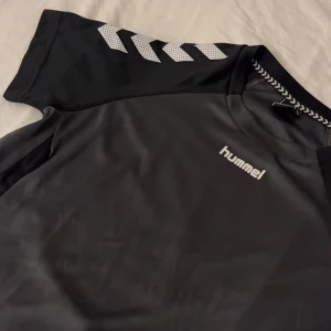 Svart/grå t-shirt från Hummel - Snygg svart t-shirt från Hummel med deras klassiska pilar på ärmen. Perfekt för sportiga aktiviteter eller en avslappnad stil. Tillverkad i ett bekvämt material som andas. Endast testad❤️❤️