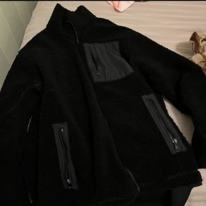 Svart teddyjacka med dragkedjor - Mysig svart teddyjacka med flera praktiska dragkedjor. Perfekt för kyliga dagar med sin fluffiga och varma design. Jackan har en hög krage och en bröstficka med dragkedja för extra förvaring.