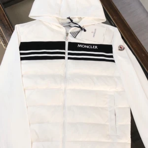Vit dunjacka från Moncler - Snygg vit dunjacka från Moncler med huva och dragkedja. Jackan har svarta ränder och Moncler-logga på bröstet. Perfekt för kyliga dagar.