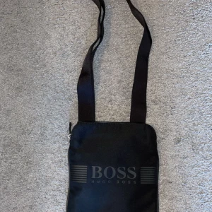Svart axelväska från Hugo Boss - Snygg svart axelväska från Hugo Boss med justerbar rem och dragkedja. Perfekt för att bära med sig det nödvändigaste med stil. Diskret logotyp på framsidan ger en elegant touch.