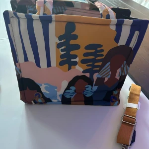 Färgglad mönstrad väska - Säljer en mellan och rymlig väska med ett unikt och färgglatt mönster i blått, rosa, brunt och gult. Väskan har en praktisk dragkedja och en justerbar axelrem för bekväm användning. Perfekt för att bära med sig allt man behöver under dagen.