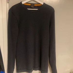 Mörkgrå tröja från BOSS - Snygg mörkgrå tröja från BOSS i slim fit. Perfekt för en stilren look med långa ärmar och enkel design. Passar bra till både jeans och chinos.