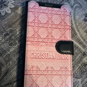 Rosa mobilfodral - Christian Dior tryck till iPhone 16 plus - Snyggt rosa mobilfodral  Christian Dior tryck  med ett geometriskt mönster och svart stängning. Fodralet har plats för kort på insidan och är perfekt för att skydda din mobil med stil.