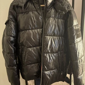 Svart pufferjacka från Michael Kors - Snygg svart pufferjacka från Michael Kors med hög krage och huva. Jackan har en glansig finish och dragkedja framtill. Nypris 2700