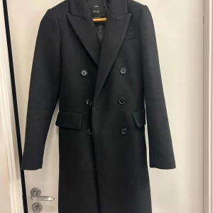 Svart rock från MNG - Elegant svart rock från MNG i ullblandning. Rocken har en klassisk design med dubbelknäppning och långa ärmar. Perfekt för kyligare dagar.