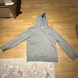 Grå hoodie från Adidas - Säljer en stilren grå hoodie från Adidas med deras klassiska logga på bröstet. Tröjan har en bekväm passform och är perfekt för vardagsbruk. Den har långa ärmar och en praktisk huva.