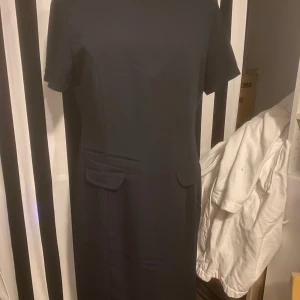 Svart klänning med korta ärmar - Elegant svart klänning med korta ärmar och en figurnära passform. Klänningen har en stilren design med diskreta fickdetaljer framtill. Perfekt för en chic och sofistikerad look.