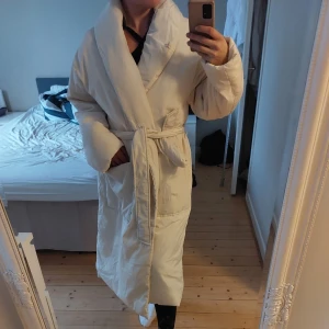 Beige kappa från Mores - Elegant quality beige kappa från Moves med knytband i midjan. Den har en lång och avslappnad passform med långa ärmar. Perfekt för kyligare dagar. Nypris 1500. Oanvänd med prislapp kvar. Storlek 42 men funkar också för dej som har 38 den blir lite oversize i modell då. Jag är en medium 
