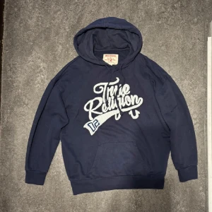 Mörkblå hoodie från True Religion - Säljer en snygg mörkblå hoodie från True Religion med stort tryck på framsidan. Perfekt för en avslappnad stil med en skön passform och huva. Märkt som strl M men skulle säga att den passar mer som S🙌🏻