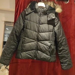 Svart quiltad vinterjacka med fuskpäls - Snygg svart quiltad vinterjacka med avtagbar huva och fuskpälsdetaljer. Jackan har dragkedja framtill och praktiska fickor med dragkedjor. Perfekt för kalla dagar. Obs trasig  på på ett ställe arm bak. Annars är den i bra skick.
