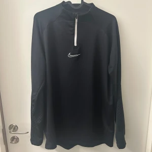 Svart långärmad tröja från Nike - Snygg och funktionell svart långärmad tröja från Nike med dragkedja och logga på bröstet. Perfekt för träning eller vardagsbruk. Tröjan har en hög krage och är tillverkad i ett mjukt material som ger komfort och stil.