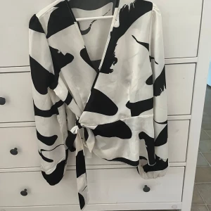 Svartvit omlottblus i polyester - Säljer en snygg svartvit omlottblus i 100% polyester. Den har ett coolt mönster och knytning i midjan. Perfekt för både vardag och fest!
