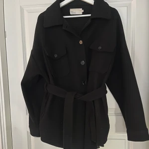 Svart skjortjacka - Oversize i storlek. Använd men i fint skick (bortsett från spännet på midjebältet som gått av, men går att byta ut/klippa av denna delen och endast knyta bältet som det är.) Midjebältet går även att ta av helt.