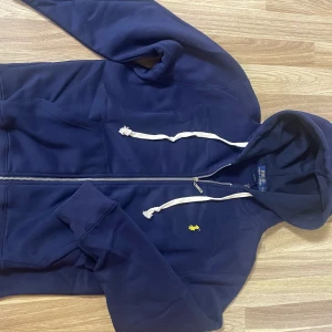 Mörkblå hoodie från Polo Ralph Lauren - Säljer en snygg mörkblå hoodie från Polo Ralph Lauren med dragkedja och justerbar huva. Den har en liten gul logga på bröstet och är perfekt för en avslappnad stil.den är använd ett par gånger och är i nyskick med väldigt skönt material.