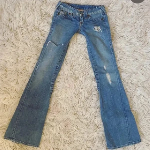Blå jeans från True Religion - Säljer ett par blå jeans från True Religion med slitna detaljer och bootcut-stil. Jeansen har en låg midja och klassiska femficksdesign med knappar på bakfickorna. Perfekta för en avslappnad look.