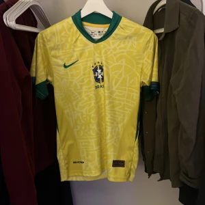 Gul Brasilien fotbollströja - Säljer en gul Brasilien fotbollströja från Nike med Neymar Jr och nummer 10 på ryggen. Tröjan har korta ärmar och gröna detaljer vid krage och ärmslut. Perfekt för fotbollsfans!