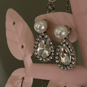 Glittriga örhängen med pärlor - Eleganta örhängen med stora glittriga stenar i droppform omgivna av mindre stenar. Örhängena har en pärla som fäste och ger en lyxig känsla. Perfekta för speciella tillfällen.