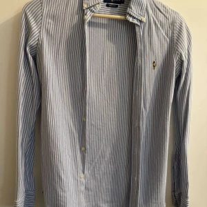 Randig skjorta från Ralph Lauren - Snygg och klassisk blå- och vitrandig skjorta från Ralph Lauren. Skjortan har långa ärmar och knappar framtill. Perfekt för en stilren look.