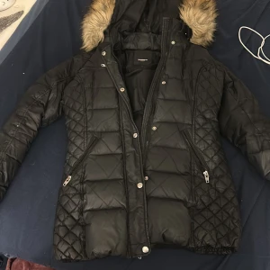 Svart dunjacka från Rockandblue - Snygg svart dunjacka från Rockandblue med quiltad design och pälskantad huva. Jackan har dragkedja och knappar framtill samt praktiska fickor med dragkedjor. Perfekt för kyliga dagar! Ett litet mini slit på armen som ej är synligt där av det mindre priset köpt för 6800 päls utbytt till fejk då någon dum sak valde att sno den äkta. Det är fejkpäls men original köpt från rockandblue.tvättas innan frakt 