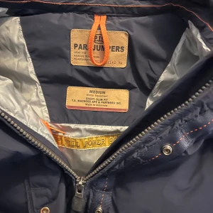 Svart vinterjacka från Parajumpers - Säljer en svart vinterjacka från Parajumpers med pälsdetaljer på huvan. Jackan har en dragkedja och knappar framtill samt en broderad logga på ärmen. Perfekt för kalla vinterdagar.