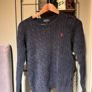 Mörkblå stickad tröja från Ralph Lauren - Säljer en snygg mörkblå stickad tröja från Ralph Lauren med det klassiska röda logotypbroderiet på bröstet. Tröjan har en kabelstickad design och långa ärmar, perfekt för kyligare dagar. Skriv om ni undrar något. Den är i storlek L14-16 men den passar xs och xxs. Pris kan diskuteras.