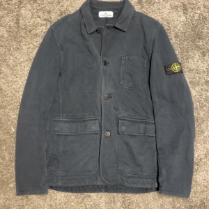 Mörkblå jacka från Stone Island Junior - Snygg mörkblå jacka från Stone Island Junior med klassisk design och knappar framtill. Jackan har en bröstficka och två stora fickor nedtill. Den ikoniska Stone Island-loggan sitter på ärmen och passar dig perfekt som är mellan 160/170