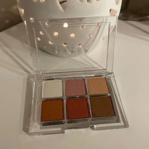 CAIA Ögonskuggspalett  - Snygg ögonskuggspalett med sex olika färger i en transparent förpackning. Innehåller nyanser av vitt, rosa, guld, brons och koppar. Perfekt för att skapa både vardagliga och festliga looks.