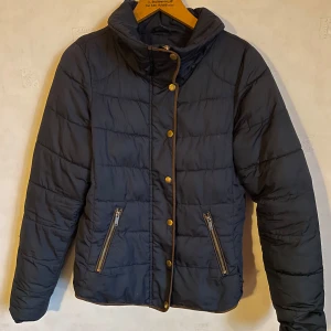 Mörkblå vinterjacka från Vila - Snygg mörkblå täckjacka från Vila med knappar och dragkedja framtill. Jackan har en hög krage och två praktiska fickor med dragkedjor. Perfekt för kyliga dagar.