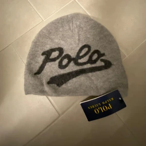 Grå mössa från Polo Ralph Lauren - Ljusgrå mössa från Polo Ralph Lauren med svart text 'Polo' (framsida) och siffrorna '1967' (baksida). 