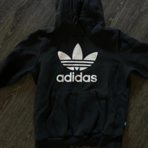 Svart hoodie från Adidas - Snygg svart hoodie från Adidas med det klassiska logotyptrycket i vitt på framsidan. Tröjan har en stor ficka framtill och en bekväm huva. Perfekt för en avslappnad stil. Passar som XS/S. 