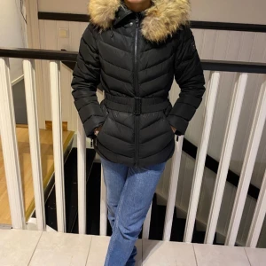 Svart dunjacka från Hollies - Snygg svart dunjacka från Hollies med pälskrage och bälte i midjan. Jackan har dragkedja och ett leopardmönstrat foder som ger en cool touch. Perfekt för kalla dagar!  (Det tillkommer även en förvarings påse till jackan då man kan hänga upp jackan med den som skydd för pälsen osv)