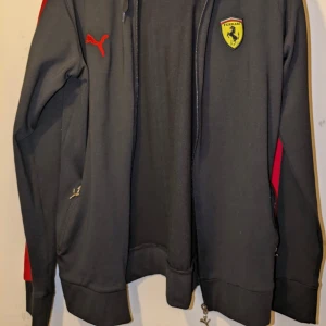 Svart jacka från Puma med Ferrari-logga - Snygg svart jacka från Puma med Ferrari-logga på bröstet. Jackan har röda detaljer och dragkedja framtill. 