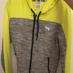 Tränings hoodie Victoria's Secret PINK - Säljer en snygg grå och gul huvtröja från Victoria's Secret PINK. Tröjan har en dragkedja framtill och en liten logotyp på bröstet. Perfekt för en avslappnad stil.