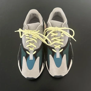 Adidas Yeezy 700 Wave Runner - Snygga Adidas Yeezy sneakers med en mix av grå, blå och svarta nyanser. Skorna har neongula snören och en unik sula med svarta och vita mönster samt orange detaljer. Perfekta för dig som vill ha en stilren men ändå iögonfallande look.
