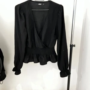 Svart omlottopp från BikBok - Snygg svart omlottopp från BikBok med långa ärmar och volangdetaljer i midjan. Perfekt för en stilren look. Toppen har en v-ringad design och är tillverkad i ett mjukt material.