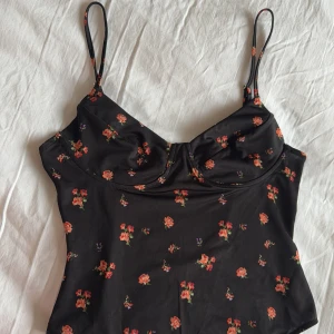 Svart blommig bodysuit - Svart bodysuit med blommigt mönster. Den har smala axelband och en öppen ryggdetalj. Strl M. Aldrig använd.