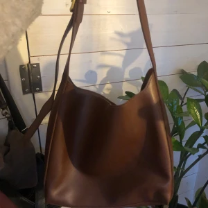 Brun axelväska i skinn - Snygg brun axelväska i skinn med enkel design. Väskan har en justerbar axelrem och stängs med magnetknapp. Perfekt  till skolan för en dator får plats