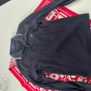 Svart långärmad pikétröja från Fred Perry - Snygg svart pikétröja från Fred Perry med klassisk krage och broderad logga på bröstet. Tröjan har långa ärmar och en stilren design med gul och vit rand på kragen.