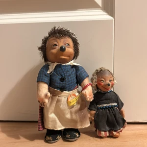 Steiff MECKI Hedgehog (igelkott) - Populära antika igelkottar från Steiff, Tyskland, klädda i trationella arbetarkläder från 1940 talet. Fin julklapp för samlare av vintageleksaker! Mamman Micki har en blå- och rutig skjorta med en beige förkläde och en röd- och svartmönstrad kjol. Dottern Mucki bär en svart klänning med ett blått band i midjan och randiga byxor.