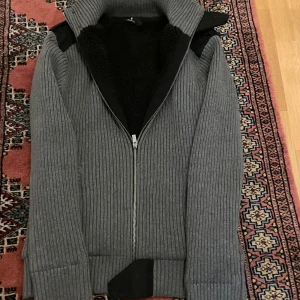 Fodrad Cardigan - Säljer en stilren grå fodrad cardigan med dragkedja och svarta axeldetaljer. Jackan har en hög krage och långa ärmar, perfekt för kyligare dagar. Den ribbade designen ger en modern look.