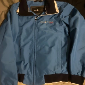 Blå jacka från Sail Racing - Snygg blå jacka från Sail Racing med dragkedja och ribbade muddar. Perfekt för vår och höst. Jackan har en stilren design med Sail Racing-logga på bröstet och ryggen.