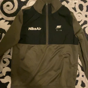 Nike tröja  - Snygg olivgrön och svart tröja från Nike Air med halv dragkedja och logga på bröstet. Perfekt för en sportig look. Tröjan har långa ärmar och en hög krage.