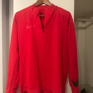 Röd långärmad tröja från Nike - Säljer en röd långärmad tröja från Nike med Dri-FIT-teknologi. Tröjan har en halv dragkedja framtill och svarta detaljer på ärmarna. Perfekt för träning och sportiga aktiviteter.