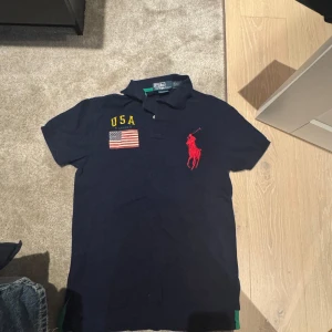 Mörkblå pikétröja från Ralph Lauren - Säljer en mörkblå pikétröja från Ralph Lauren med USA-flagga och gul text på bröstet. Tröjan har en röd broderad logotyp och korta ärmar.