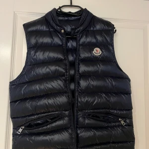 Mörkblå dunväst från Moncler - Snygg svart dunväst från Moncler med dragkedja och hög krage. Perfekt för kyligare dagar och har en stilren design med Moncler-logga på bröstet. Två praktiska fickor med dragkedja framtill. Inte äkta så kom med ett pris!